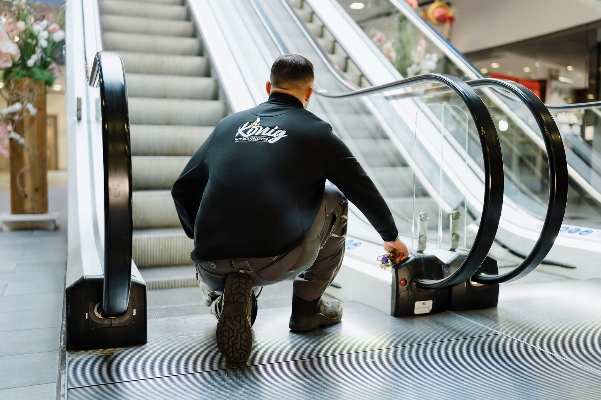 Ein Mitarbeiter des Hausmeisterservice Sven König in gebrandeter Arbeitsjacke bei der technischen Objektkontrolle an einer Rolltreppe. Die Szene veranschaulicht die proaktive und gewissenhafte Betreuung von Gewerbeimmobilien.