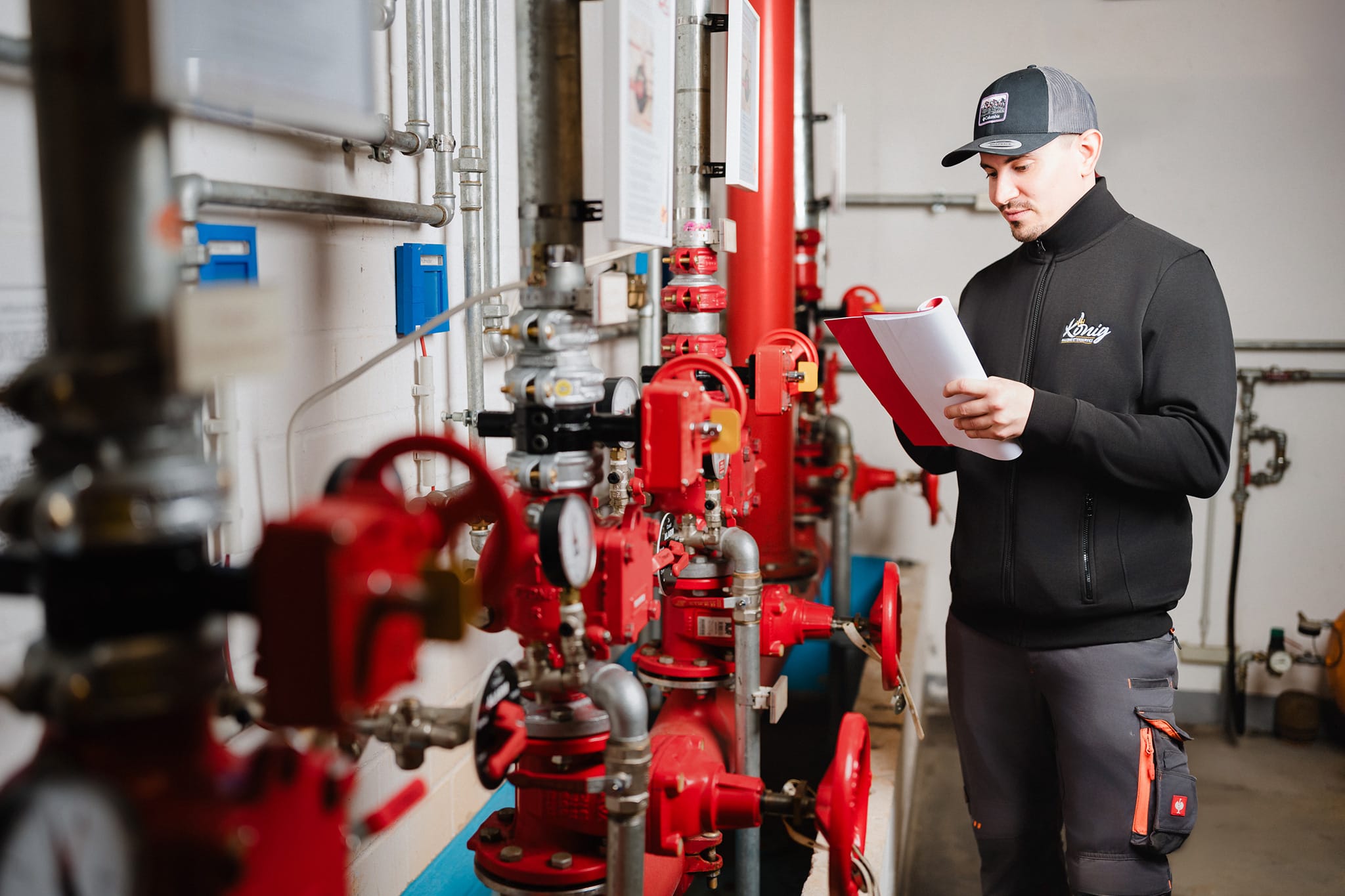 Mitarbeiter vom Hausmeisterservice Sven König in professioneller Arbeitskleidung prüft Unterlagen an einer roten Rohranlage in einem Technikraum.