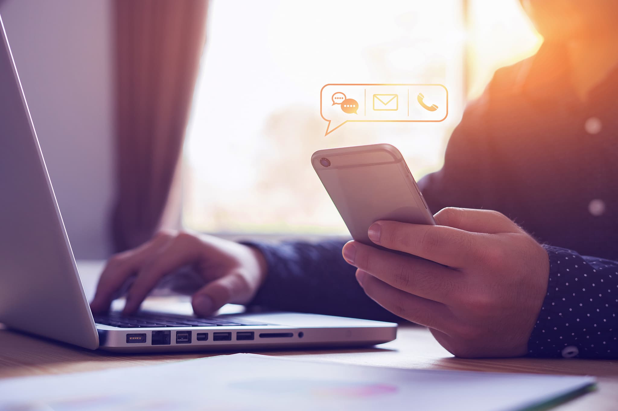 Person sitzt am Laptop und hält ein Smartphone in der Hand, über dem digitale Symbole für Nachrichten, E-Mail und Telefon schweben. Symbolbild für die schnelle Erreichbarkeit beim Hausmeisterservice Sven König in Troisdorf, Köln und Bonn.
